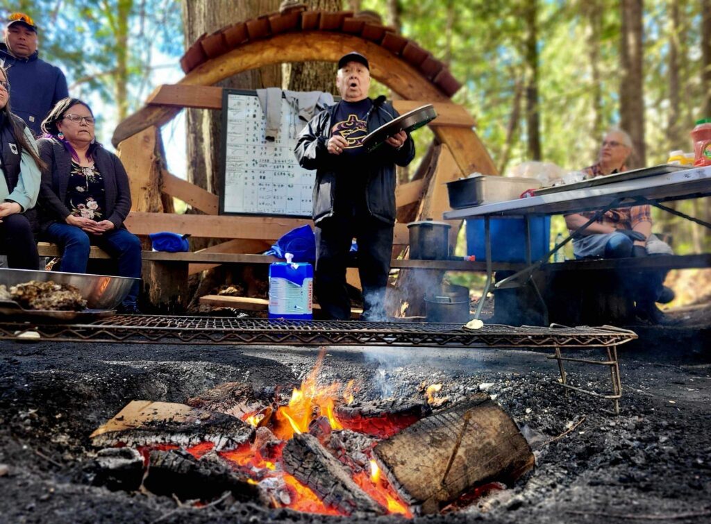 Elder George Harris is drumming with a group by a fire pit at a gathering spot on the Stz'uminus trail project funded by the First Peoples' Cultural Council Heritage Program.