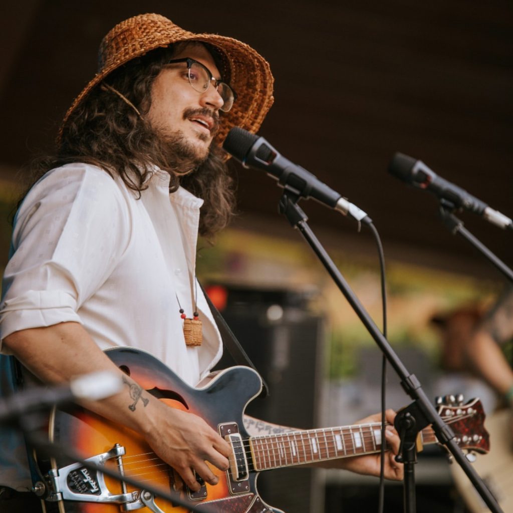 Saltwater Hank playing a guitar and wearing a woven cedar hat. His music is funded by FPCC's Indigenous music grant