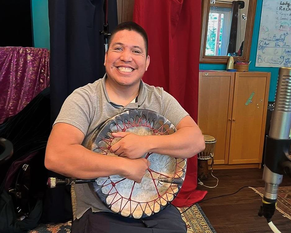 First Nations singer and songwriter Patrick Aleck smiling and holding his drum while sitting at a recording microphone.