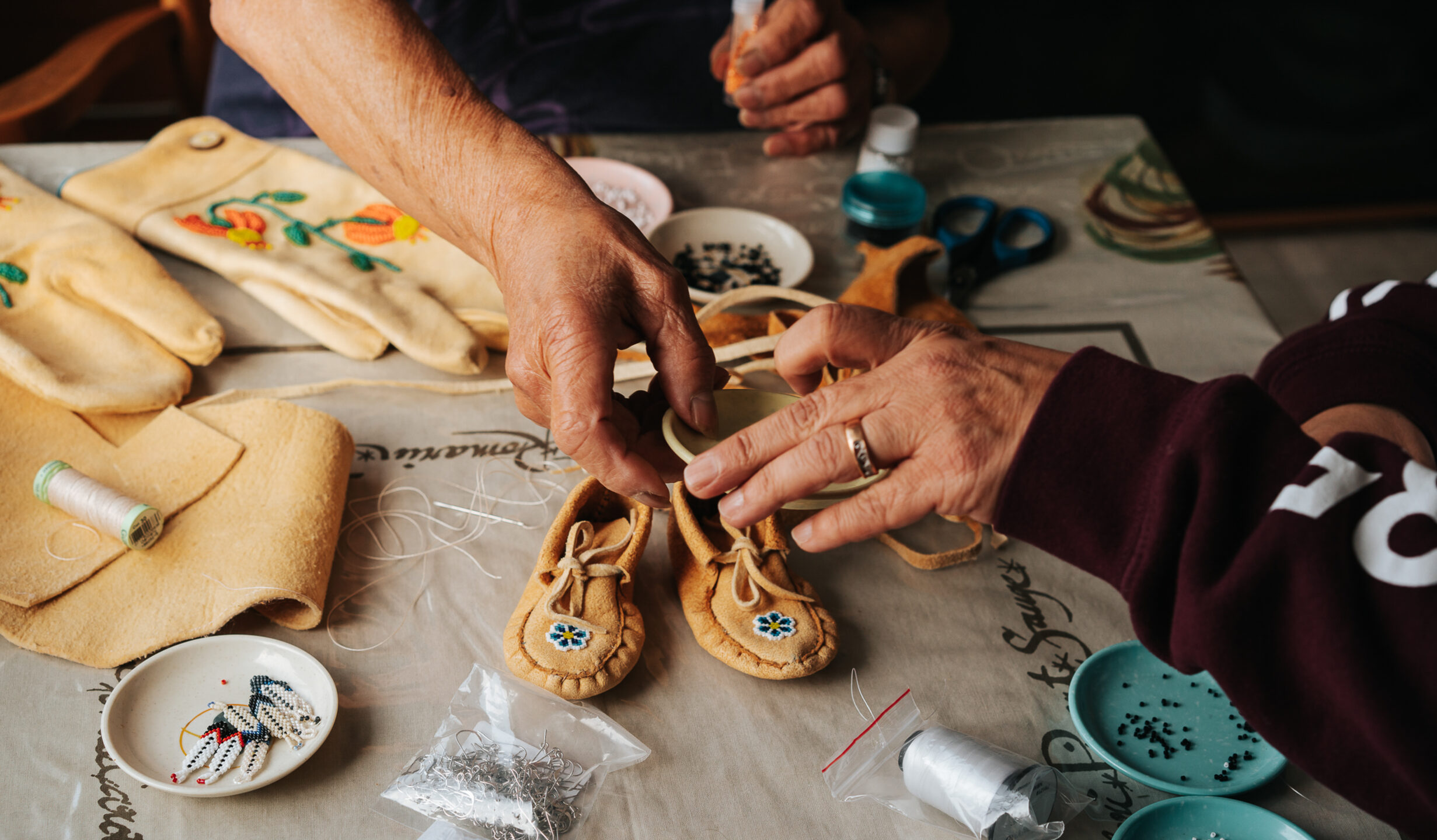The hands of two people engaged in First Nations beadwork at a table in WIliams Lake B.C. Supported by B.C. First Nations Arts Program funding at the First Peoples' Cultural Council