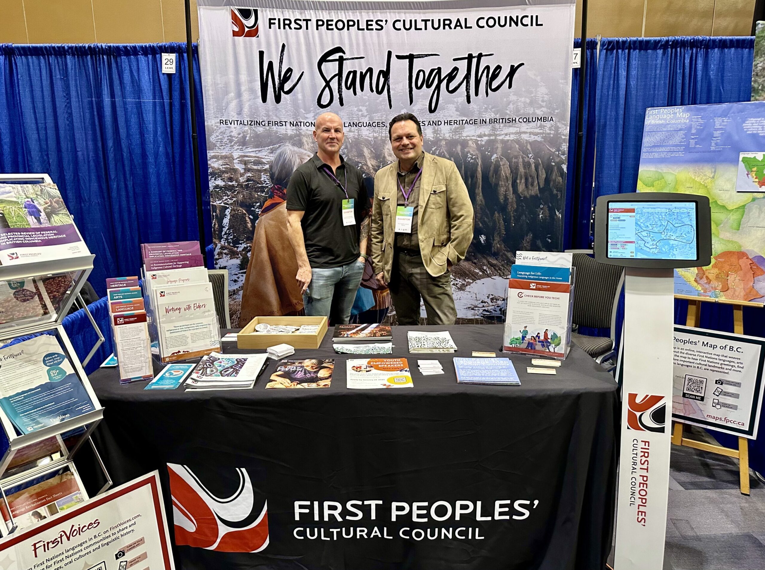 FPCC Board Members Jamie Sterritt and Gerald Lawson standing in front of the trade show booth for the First Peoples' Cultural Council at the BC Cabinet and First Nations Leadership Gathering in 2023.
