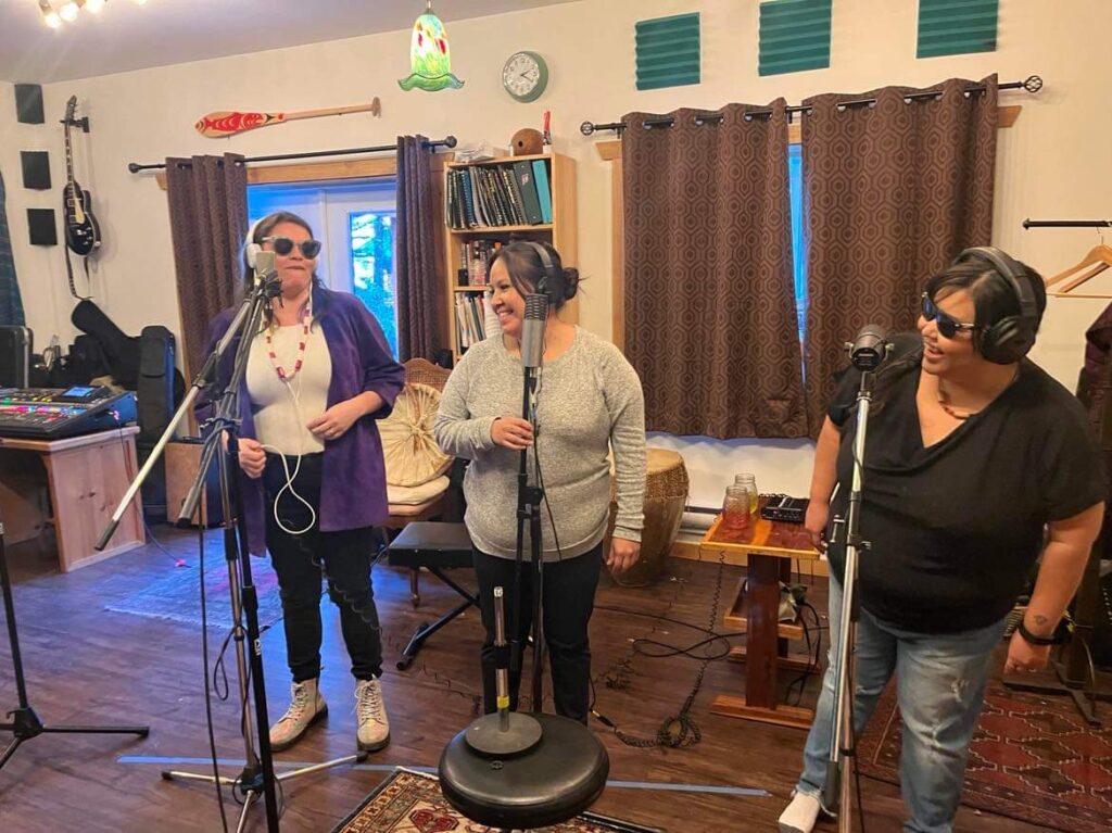 Three First Nations women stand smiling and laughing at microphones and wearing headphones while recording music for an album by Patrick Aleck in Nanaimo B.C.