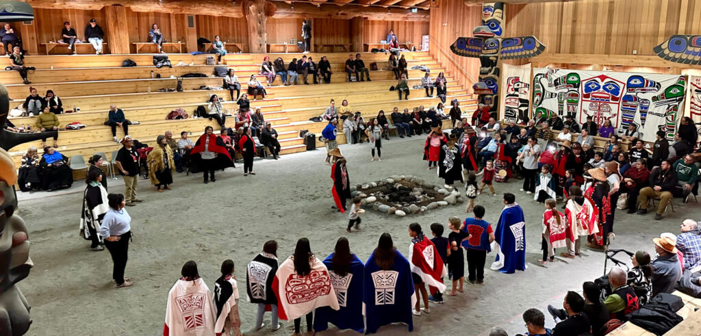 A ceremony in the Big House in Bella Bella shows the inside with large carved totems at one end. Adults and children wearing blanket in a circle on the floor dancing. Little children carry drums.