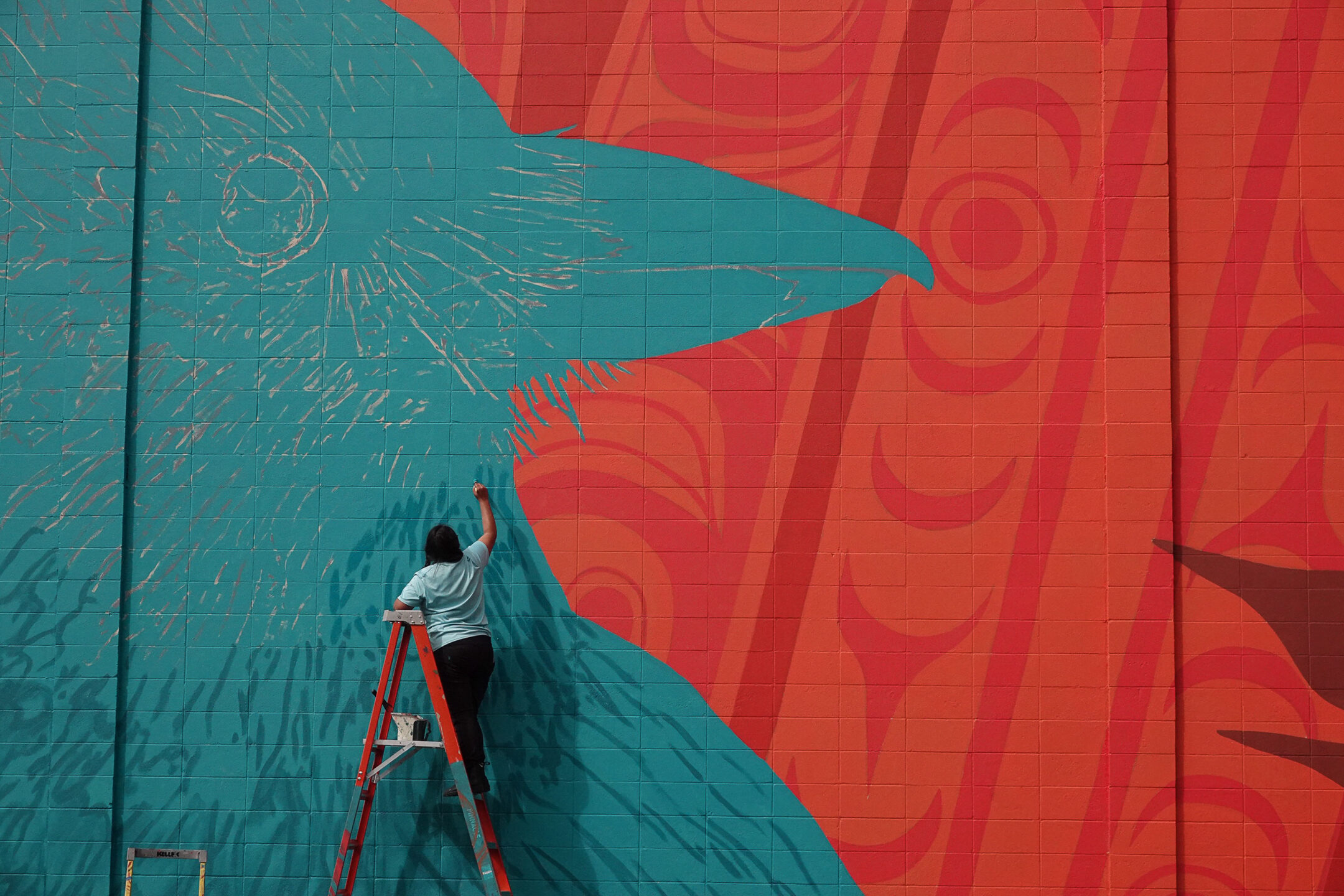 A person on a ladder paints a turquoise and red mural with a raven. Image is used to represent the FPCC Arts Vitality Grant.